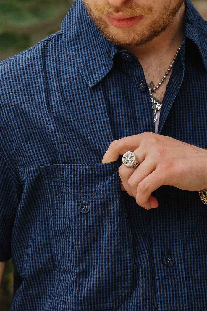Close-up of a man wearing a dark blue and black micro-check patterned short-sleeve button-up shirt, with his hand in his pocket and a silver ring on his finger.