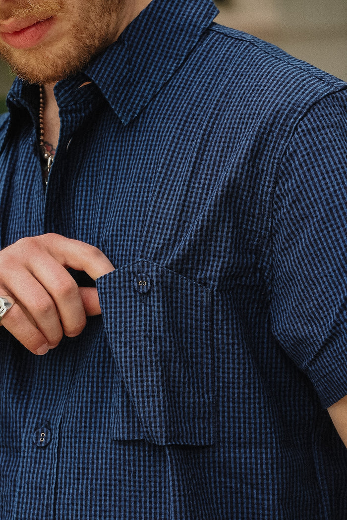 Close-up of a man wearing a short-sleeve button-up shirt with a small blue and white gingham pattern, adjusting the pocket flap.