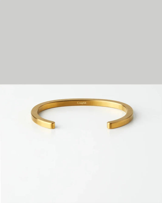 Minimalist gold metal bracelet with open cuff design on white surface, engraved Craighill logo